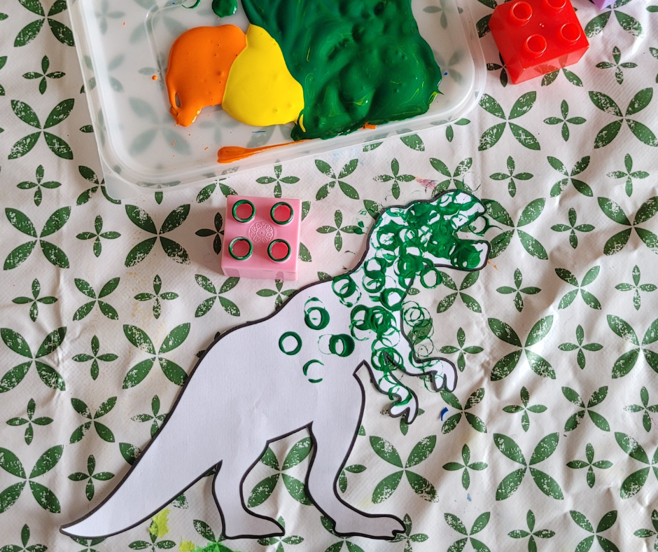 An image showing an in process photo of duplo stamped dinosaur art. There is a cut out dinosaur template that is half painted using a duplo block that is sat next to it with green paint on it, there are other duplo blocks and a tray with some paint next to it.
